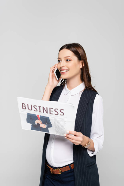 happy young pregnant businesswoman holding business newspaper and talking on smartphone isolated on grey - Valokuva, kuva