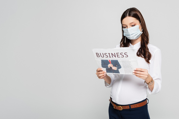 mladá těhotná podnikatelka v lékařské masce s obchodní noviny izolované na šedé - Fotografie, Obrázek