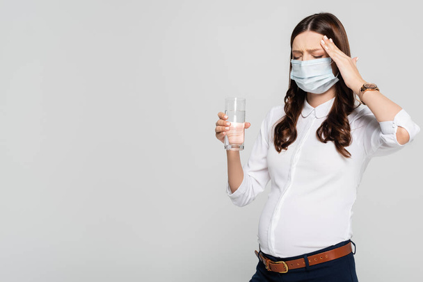 joven embarazada mujer de negocios en máscara médica con agua tener dolor de cabeza aislado en gris - Foto, imagen