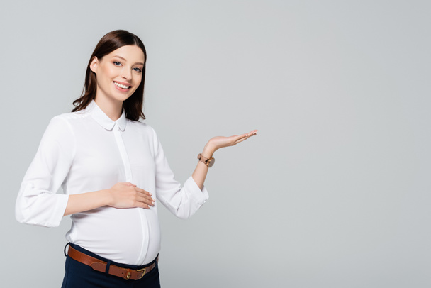 sonriente joven embarazada mujer de negocios señalando a un lado aislado en gris - Foto, imagen