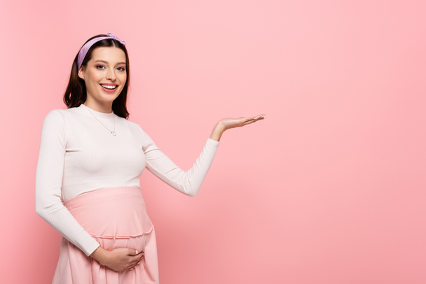 happy young pretty pregnant woman touching belly and pointing aside isolated on pink - Photo, Image