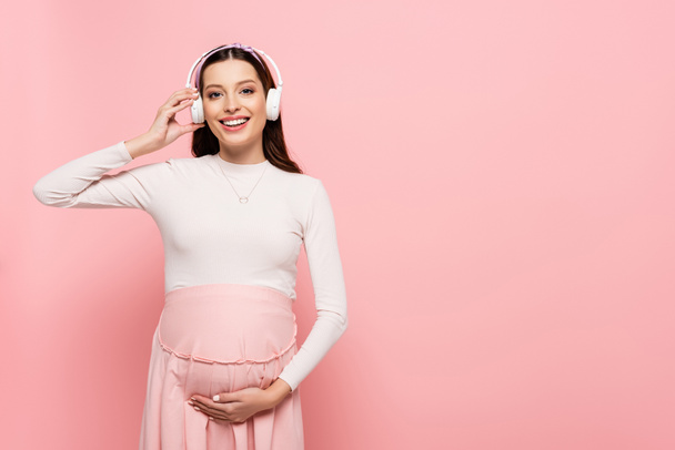 happy young pretty pregnant woman in headphones touching belly isolated on pink - Foto, afbeelding