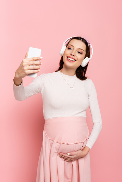 счастливая молодая красивая беременная женщина в наушниках, используя смартфон изолирован на розовый - Фото, изображение