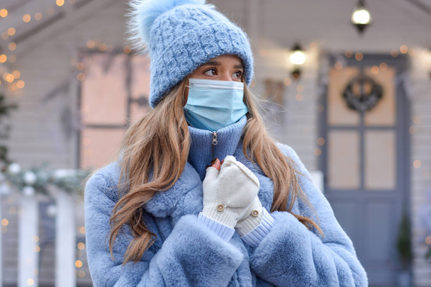 Een mooi meisje, model, in een hoed, bontjas, handschoenen, draagt een medisch masker buiten tegen de achtergrond van een kersthuis. Kerst veilig ontmoeten tijdens de coronavirusuitbraak. - Foto, afbeelding