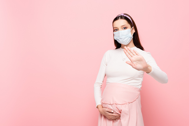 joven embarazada en máscara médica mostrando gesto de stop aislado en rosa - Foto, Imagen