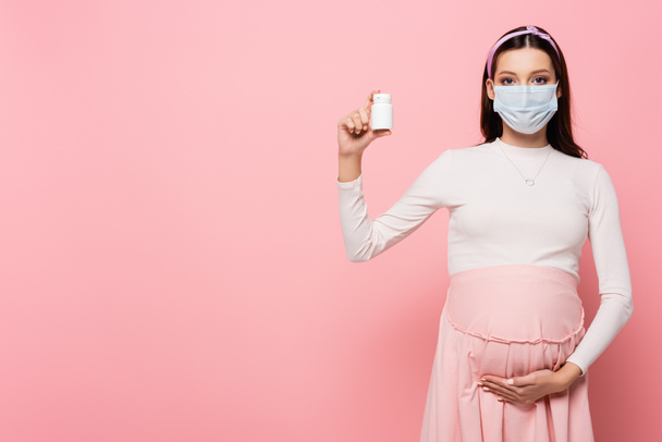 νεαρή έγκυος γυναίκα σε ιατρική μάσκα με χάπια που απομονώνονται σε ροζ - Φωτογραφία, εικόνα