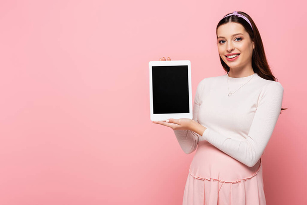 feliz joven bonita mujer embarazada con tableta digital aislada en rosa - Foto, Imagen