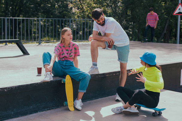 An einem schönen sonnigen Tag, Gruppe von Freunden sehr attraktiv, multiethnisch genießen die Zeit in einem modernen Skatepark, sie sitzen auf dem Boden und Geselligkeit vor dem Start zum Skateboarden - Foto, Bild