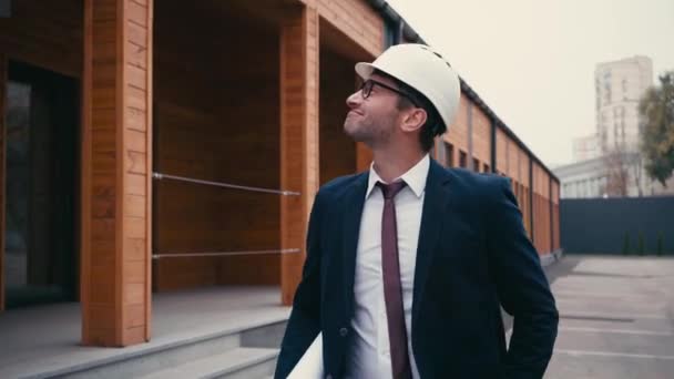 χαρούμενος αρχιτέκτονας με κοστούμι και hardhat χαμογελώντας, ενώ το περπάτημα κοντά στο κτίριο - Πλάνα, βίντεο