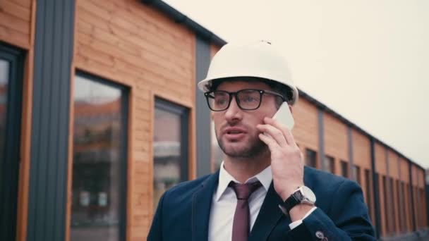 arquitecto disgustado en traje y hardhat hablando en teléfono inteligente cerca del edificio - Metraje, vídeo