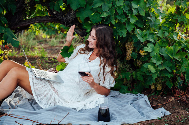 Beautiful woman in white dress works with laptop online and drinks wine - Фото, изображение
