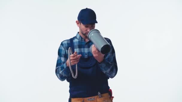 смущенный сантехник в униформе, держащий шланг и трубу изолированы на белом - Кадры, видео