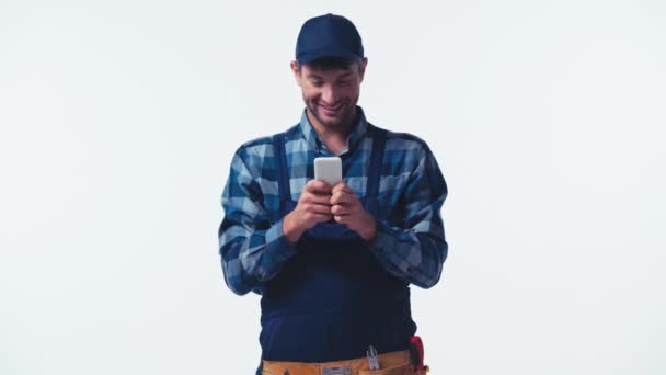 šťastný instalatér v uniformě a čepice textování na smartphone izolované na bílém - Záběry, video