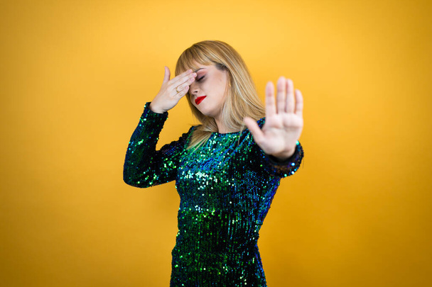 Beautiful blonde woman wearing a fancy dress over yellow background covering eyes with hands and doing stop gesture with sad and fear expression. Embarrassed and negative concept. - Photo, Image