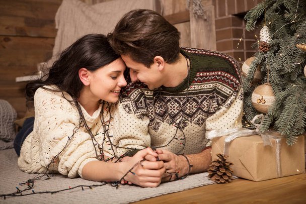 Beautiful brunette caucasian romantic loving couple in cozy warm sweaters in the cabin on a christmas new year eve, smiling, kissing, having fun with each other. Dim dark night light, toned image - Photo, Image