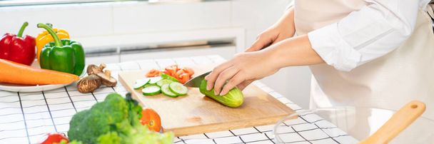 νεαρή Ασιάτισσα γυναίκα ετοιμάζει σαλάτα λαχανικών υγιεινής διατροφής κόβοντας αγγούρι για τα συστατικά στο ξύλο κοπής σε ελαφριά κουζίνα, μαγείρεμα στο σπίτι και την έννοια των υγιεινών τροφίμων - Φωτογραφία, εικόνα