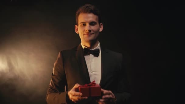 Focus pull of smiling young man in suit showing red gift box on black background - Footage, Video