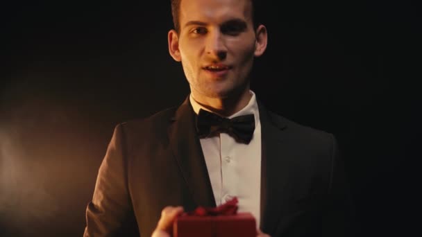 Rack focus of happy man in suit talking and showing present on black background - Footage, Video