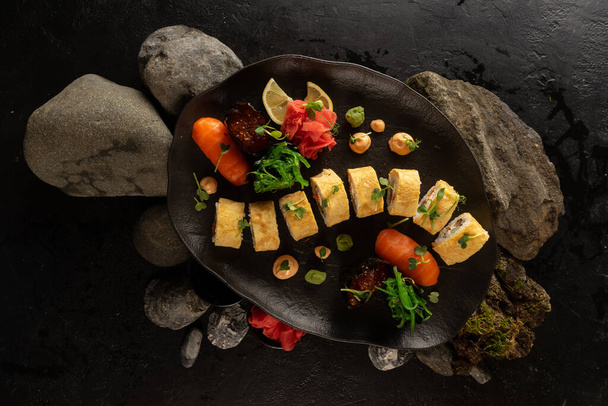 Un juego de sushi en un hermoso plato negro. Plato japonés. Rollos de sushi con amlet de tamago japonés y sashimi de salmón con arroz, gunkans con caviar rojo, jengibre, wasabi, ensalada de chuka y salsa de maní. - Foto, imagen