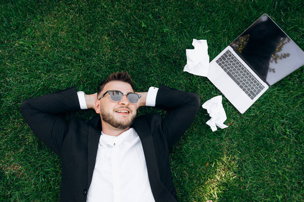 芝生の上でリラックス。草の上に寝そべっている間仕事から休んでいるスタイリッシュな正式なスーツとサングラスを身に着けている確信している白人ビジネスマン。近くにノートパソコンがある。トップ表示 - 写真・画像
