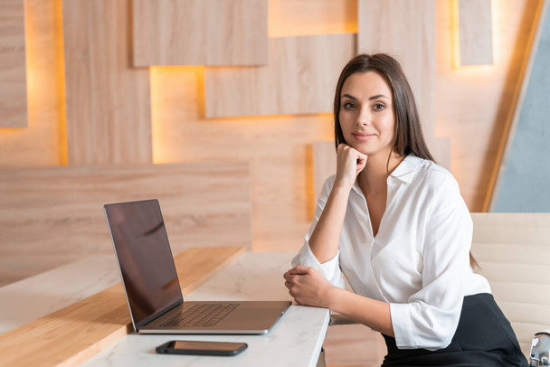 Office manager woman in white shirt, beautiful smile, sitting to table with laptop, hand to chin, in wooden office interior. Friendly office worker looking at the camera. Concept of work - Foto, Imagem