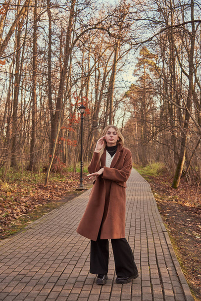 A blonde charming girl in a brown coat walks along an alley in an autumn Park. - Fotó, kép