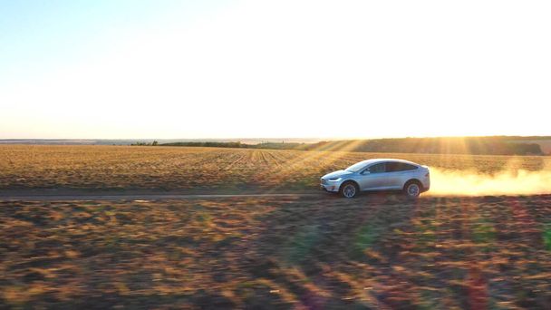 電気自動車の空中ショットオフに移動する道路ルートの背後にほこり歩道を残します。農村部の道路に沿って電荷駆動に生態に優しい自動車。背景に風景夕日の風景です。サイドビュー. - 写真・画像