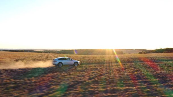 Aerial shot of electrical car moving on off-road route leaving dust trail behind. Ecology friendly auto on electric charge driving along rural road. Scenic sunset landscape at background. Side view. - Photo, Image