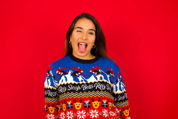 fiatal gyönyörű kaukázusi nő visel karácsonyi pulóver piros fal, boldog és vicces arc mosolygó és bemutató nyelv. - Fotó, kép