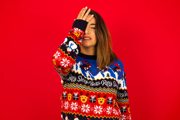 Разочарованная белая женщина в рождественском свитере, прислоненном к красной стене, держась за руку на лбу в депрессии, сожалея о том, что он сделал, у него была головная боль, он выглядел напряженным. - Фото, изображение