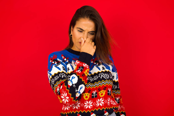 mujer caucásica vistiendo suéter de Navidad contra la pared roja sosteniendo su nariz debido a un mal olor. - Foto, Imagen