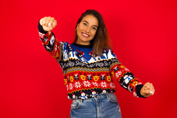 Retrato de una encantadora mujer caucásica usando suéter de Navidad contra la pared roja, sonriendo ampliamente mientras toma las manos sobre su cabeza. Concepto de confianza y relajación. - Foto, Imagen