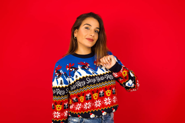 Крупный план веселой молодой красивой белой женщины в рождественском свитере у красной стены, выглядит радостным, довольным и уверенным, указывает на себя большим пальцем. - Фото, изображение