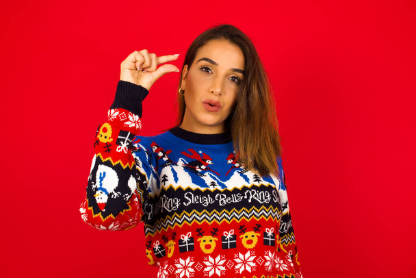 Шокированная молодая красивая белая женщина в рождественском свитере, прислоненном к красной стене, показывает что-то малое руками, демонстрирует размер, открывает рот от удивления. Концепция измерения. - Фото, изображение