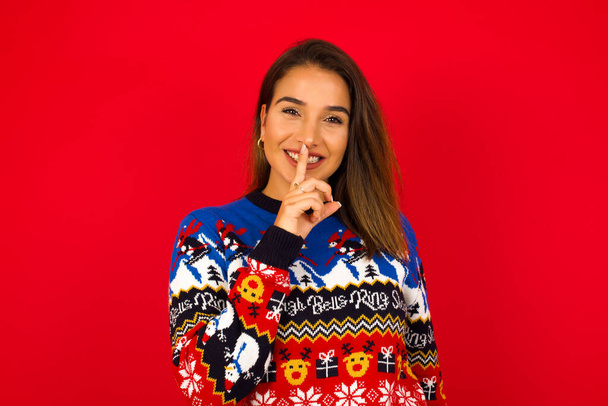 Smiling Young beautiful Caucasian woman wearing Christmas sweater against red wall,  makes shush gesture, holds fore finger over lips hides secret. Be mute, please. - Photo, Image