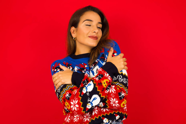 Junge schöne kaukasische Frau im Weihnachtspullover vor roter Wand, sich glücklich und positiv umarmt, selbstbewusst lächelnd. Selbstliebe und Selbstfürsorge. - Foto, Bild