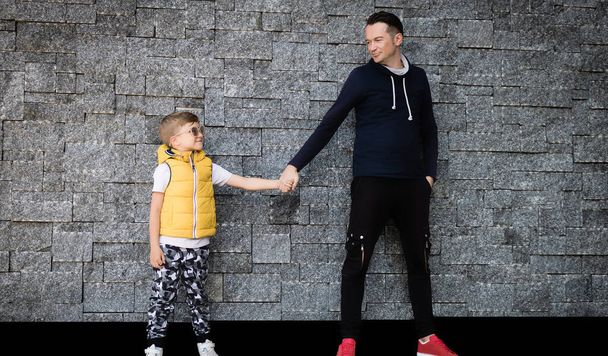 Gülümseyen çocuk duvara karşı ayakta dururken babasıyla el ele tutuşuyor..  - Fotoğraf, Görsel