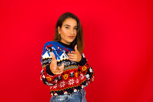 Гей, ти, бац. Радісна і харизматична вродлива молода кавказька жінка, одягнена у різдвяний светр проти червоної стіни, підморгує пальцями на камеру, щасливо і нахабно.. - Фото, зображення