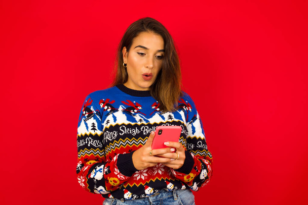 若い美しい白人女性が赤い壁にクリスマスセーターを着て、盗聴された目で見て、現代のスマートフォンを保持し、友人から予期しないメッセージを受け取り、リマインダーを読む. - 写真・画像
