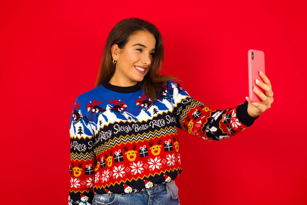 Kırmızı duvara karşı Noel süveteri giyen mutlu, genç, beyaz bir kadın cep telefonuyla selfie çekiyor. İnsanlar, teknoloji ve eğlence kavramı - Fotoğraf, Görsel