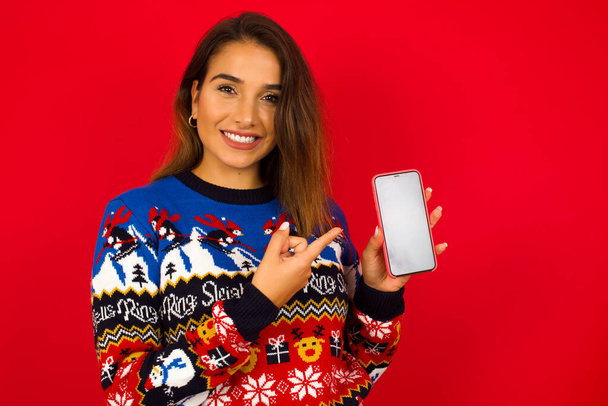 Sonriente Joven hermosa mujer caucásica con suéter de Navidad contra la pared roja, mostrando y apuntando a la pantalla del teléfono vacía. Concepto de publicidad y comunicación. - Foto, imagen