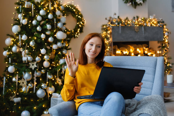 クリスマスの挨拶をオンラインで。笑顔の女性は友人や両親へのビデオ通話を行うためにコンピュータ、ラップトップを使用しています。クリスマスツリー、暖炉、ライトガーランド付きの居心地の良いリビングルームに座って - 写真・画像