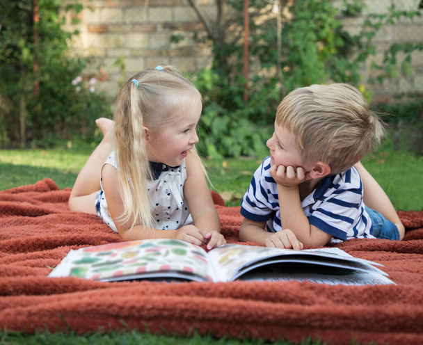 Cute blond toddler boy and girl lie on their bellies on soft brown blanket in summer on lawn. they look at each other cheerfully with interest, before them an open book. Useful entertainment - Photo, Image
