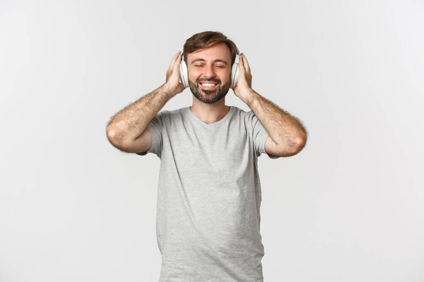 Imagen del hombre guapo en traje casual, escuchando música en auriculares inalámbricos, sonriendo complacido, de pie sobre fondo blanco - Foto, Imagen