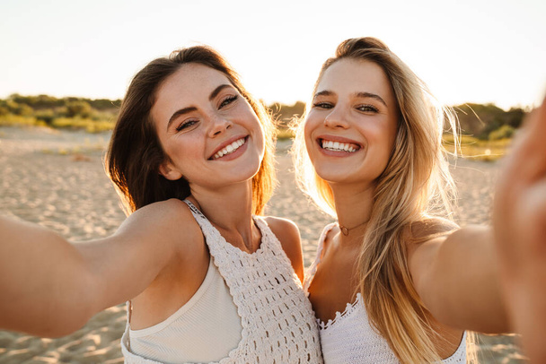 Two young caucasian happy women smiling and taking selfie photo while walking on beach - Foto, Imagen