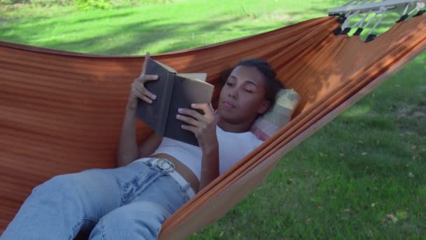 Młoda kobieta czyta książkę leżąc w hamaku w lesie - Materiał filmowy, wideo