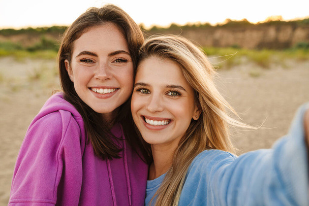 Two young caucasian happy women smiling and taking selfie photo while walking on beach - Foto, Imagem