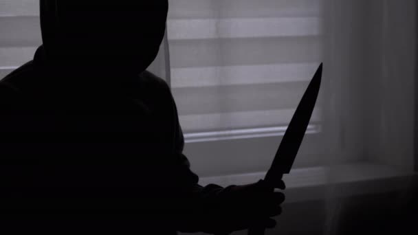 Silueta de niño solitario en capucha con cuchillo en la mano cerca de la ventana. Asesino adolescente - Metraje, vídeo