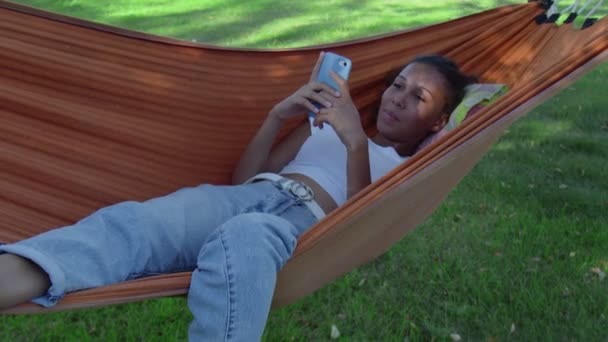 Молодая африканская американка с мобильным телефоном отдыхает на гамаке. - Кадры, видео