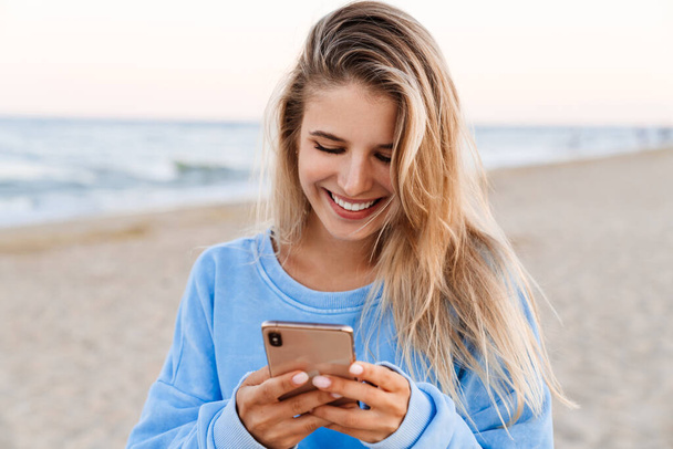 Young caucasian blonde woman smiling and holding cellphone while walking outdoors - Foto, Imagem